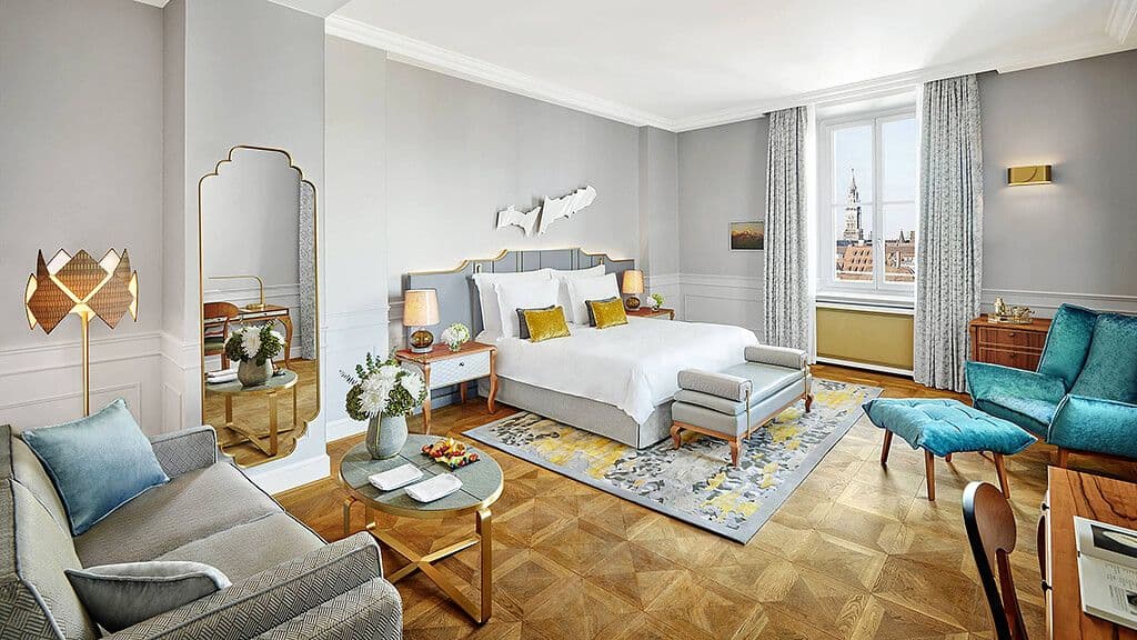 A hotel room with a bed, a seating area, and a window looking out over Munich rooftops