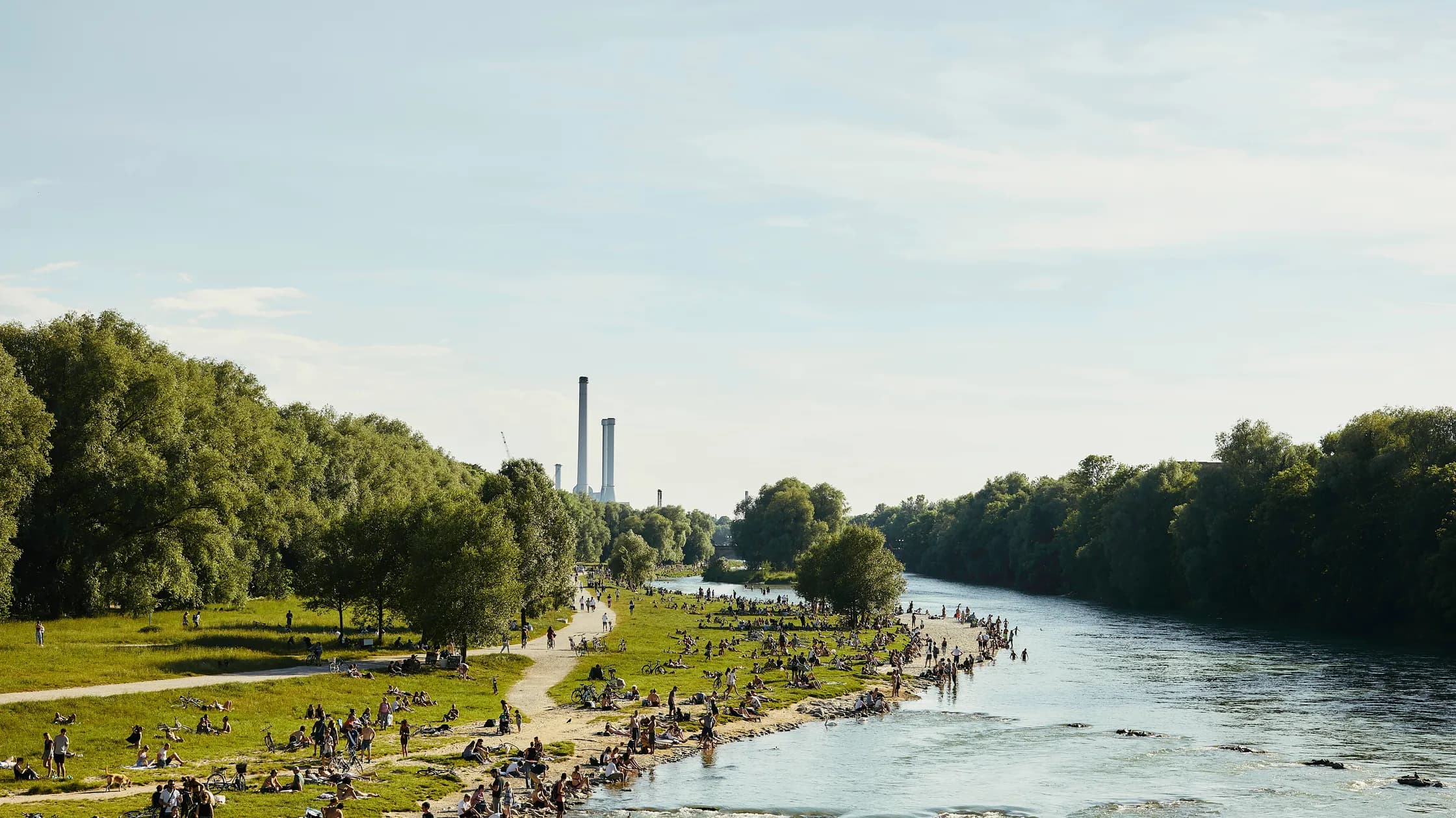 Crowds sunbathing and walking along the gravel banks of the Isar river in Munich, with cooling towers visible in the distance