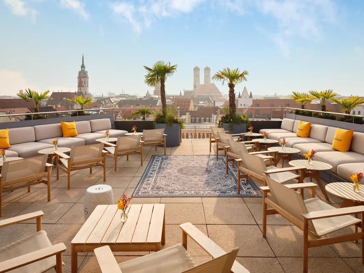 A hotel rooftop terrace with lounge seating and palm trees, the Frauenkirche's towers on the skyline behind